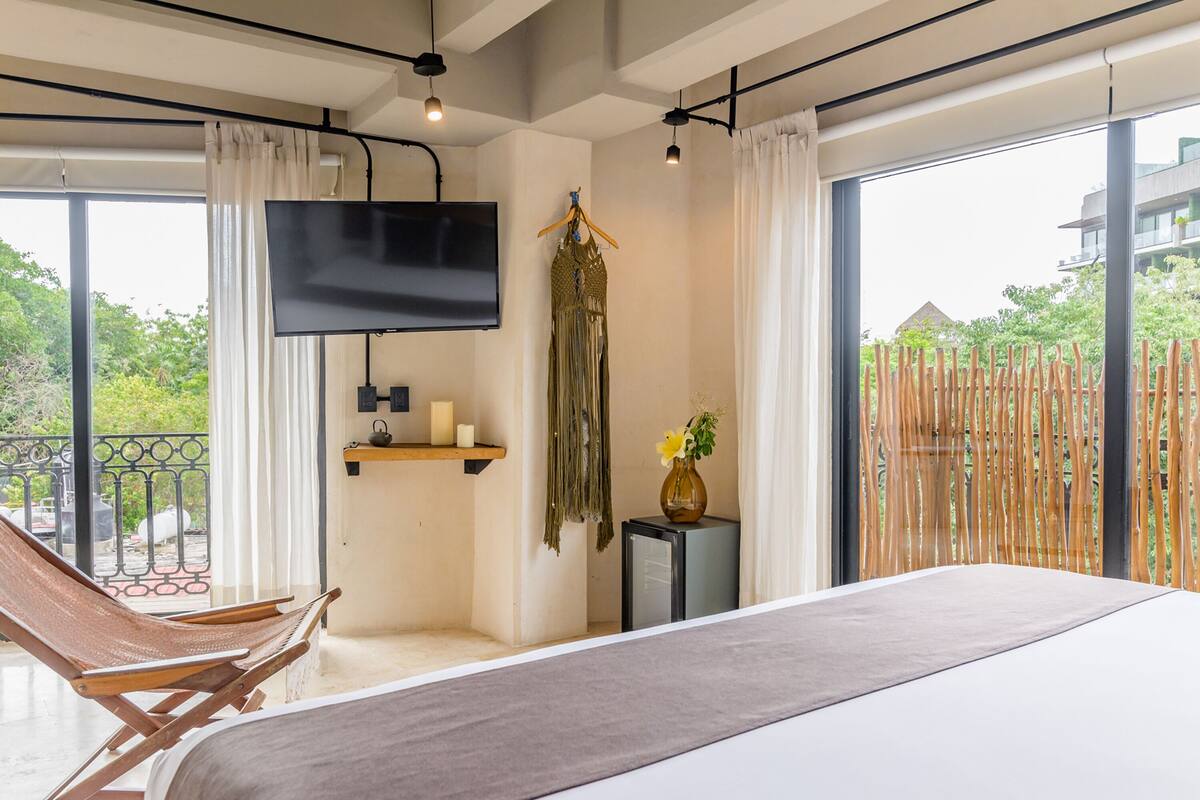 A modern hotel room with a large bed, a TV mounted on the wall, a wooden chair, and a balcony with a view of greenery.
