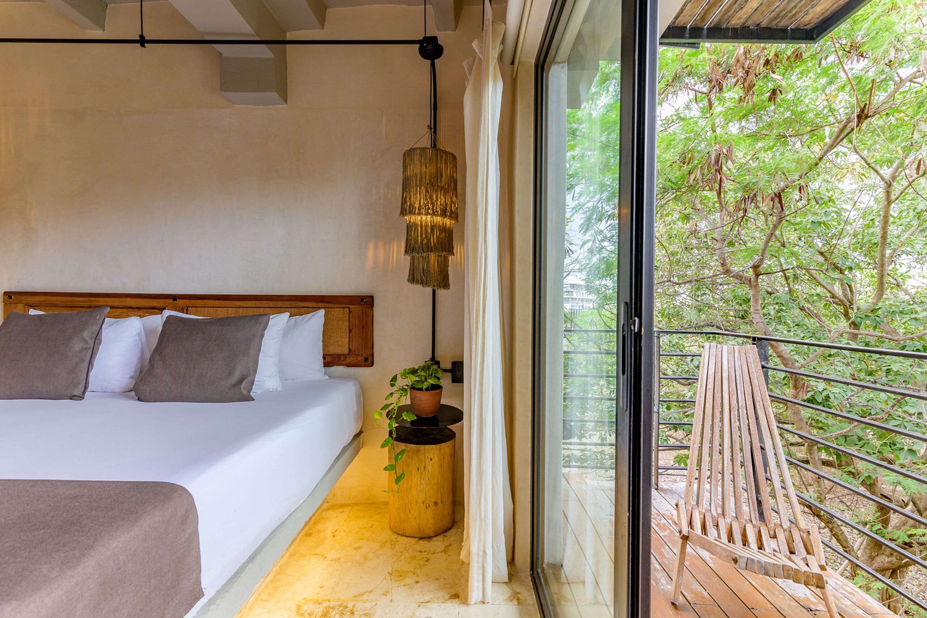 Un dormitorio con una cama, un lámpara colgante, una planta en maceta y un balcón con una silla de madera.