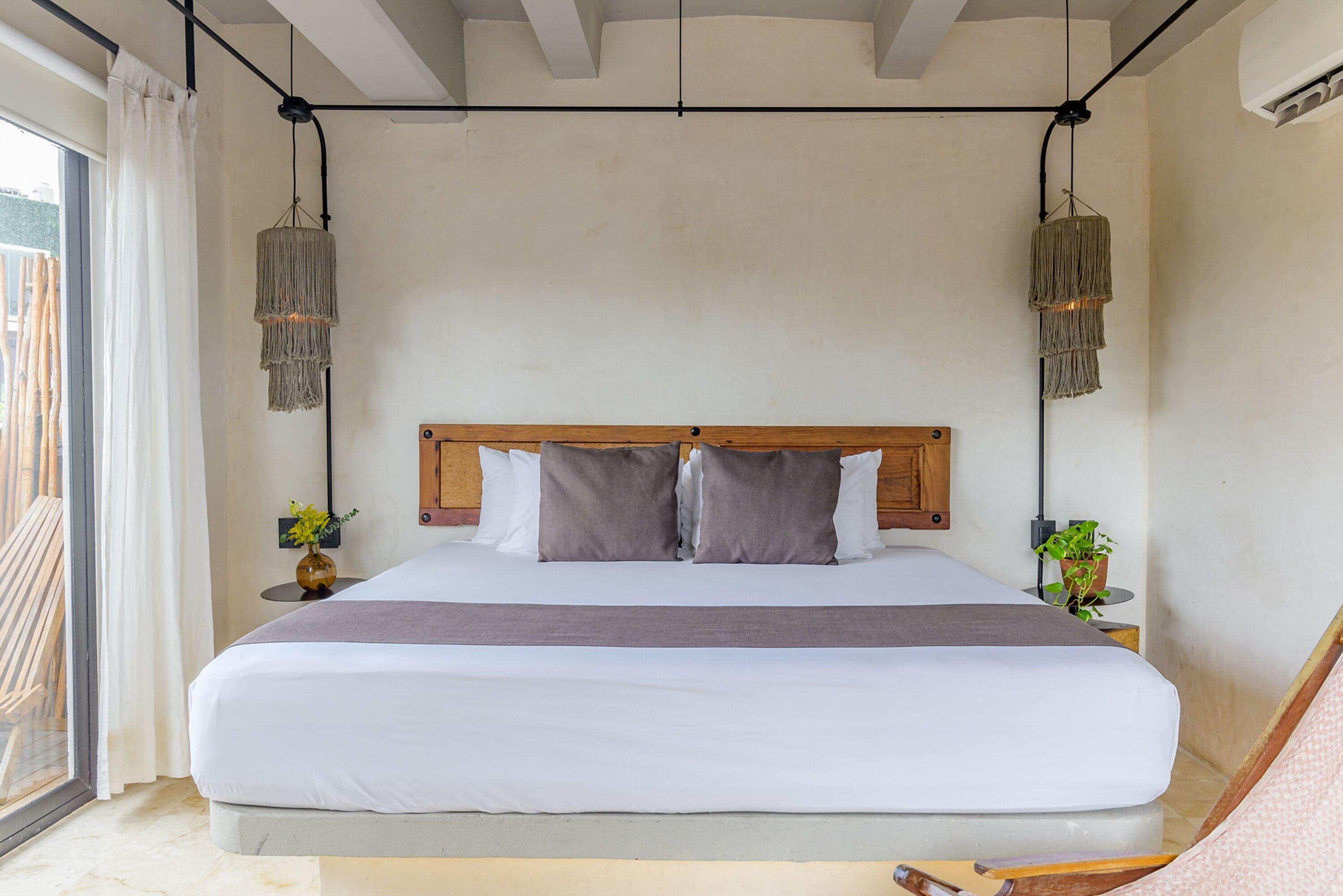A large bed with a wooden headboard, two bedside tables with plants, and a view of the outside through a sliding door.