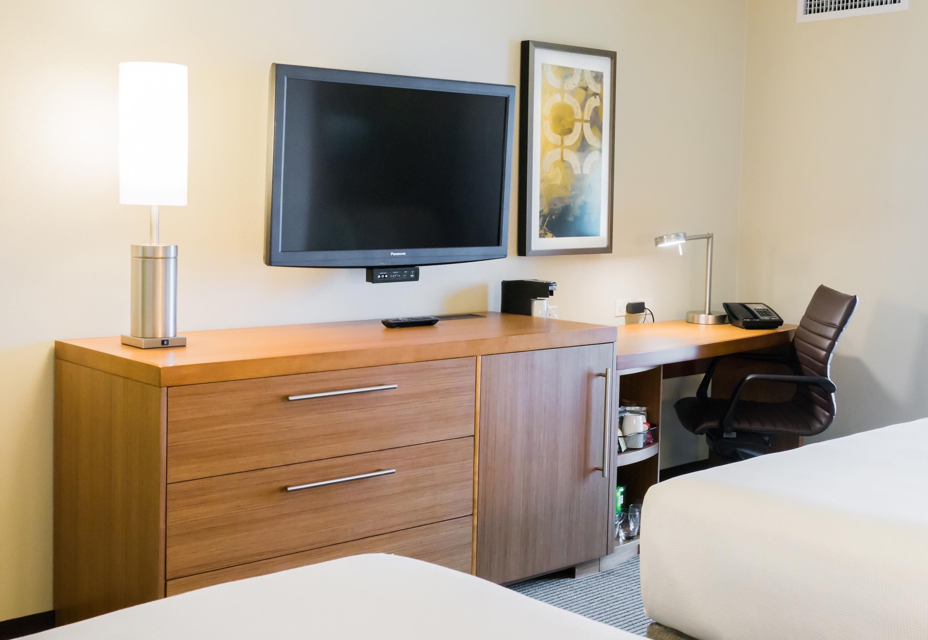 A hotel room with a bed, a desk with a flat-screen TV, a lamp, a chair, and a cabinet.