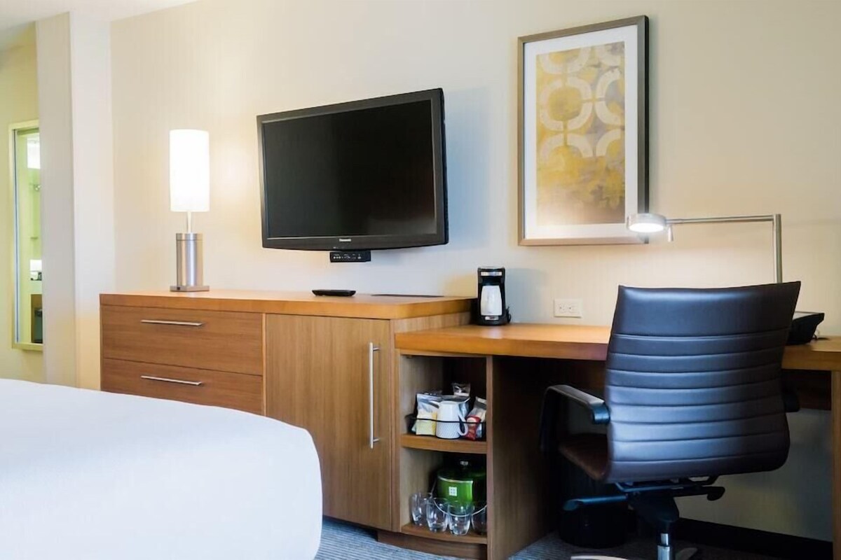 A hotel room with a bed, a desk with a chair, a television, a lamp, a coffee maker, and a shelf with kitchen items.