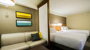A hotel room with a sofa, two beds, a nightstand, and a wall-mounted lamp.