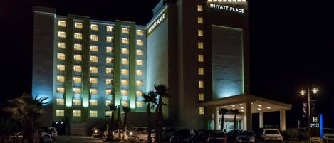 Un hotel de varios pisos con un llamativo letrero de 'Hyatt Place', un estacionamiento con varios autos y palmeras al frente.