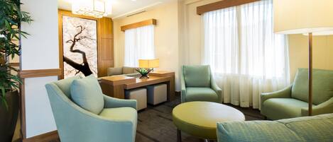 Una habitación de hotel moderna con zona de estar que incluye dos sillones y una mesa redonda, un escritorio de madera y un amplio ventanal con cortinas traslúcidas.