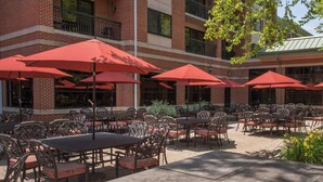 An outdoor patio with tables and chairs, red umbrellas, and a brick building with multiple windows and balconies.