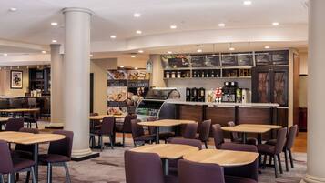 A modern restaurant with round tables and purple chairs, a bar area with a menu, and a coffee machine.