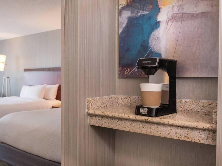 A hotel room with a coffee machine on a granite countertop, a painting on the wall, and a bed with a headboard.