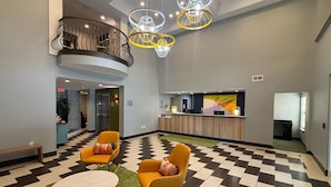 Un hall d’entrée avec un sol à carreaux, deux chaises jaunes, une table ronde, un comptoir d’accueil et un escalier.