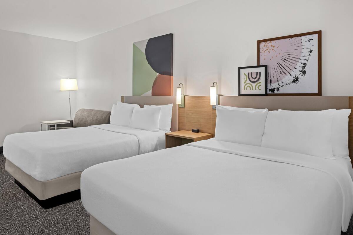 A hotel room with two beds, a nightstand, a lamp, and two wall-mounted light fixtures.