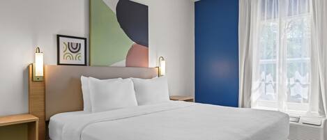A hotel room with a large bed, two bedside tables, a wall art piece, a framed picture, and a window with sheer curtains.