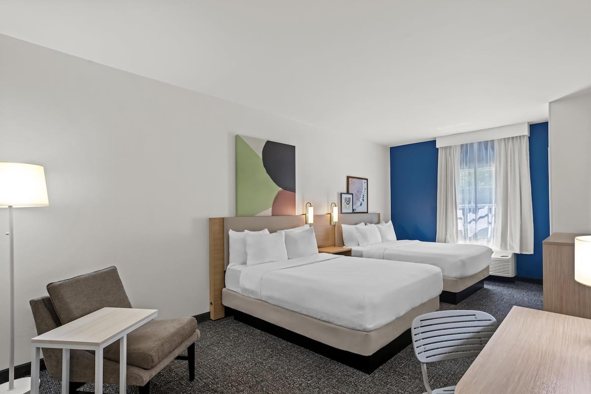 A hotel room with two beds, a desk, a chair, and a lamp.