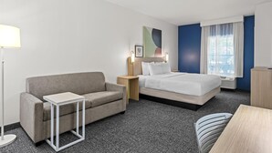 Une chambre d’hôtel avec un canapé gris, un lit avec une literie blanche, une petite table, une lampe et un tableau au mur.