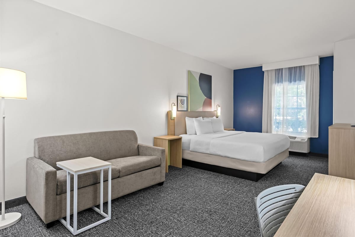 A hotel room with a grey sofa, a bed with white bedding, a small table, a lamp, and a picture on the wall.