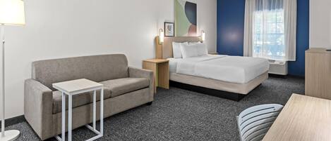 Habitación de hotel con sofá gris, cama con ropa de cama blanca, una mesita, lámpara y un cuadro en la pared.