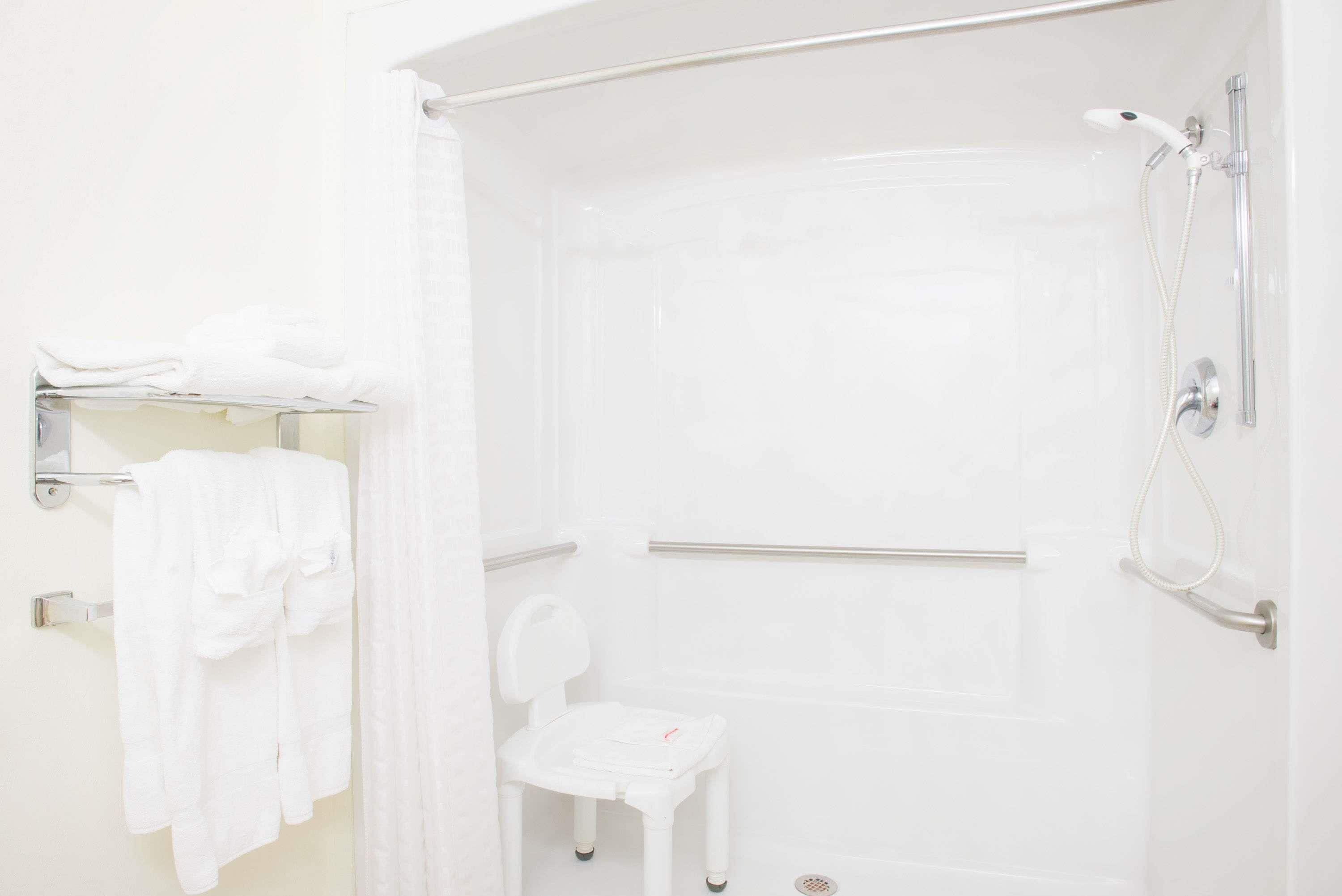 A white bathroom with a shower, a small white chair, and a towel rack with towels.