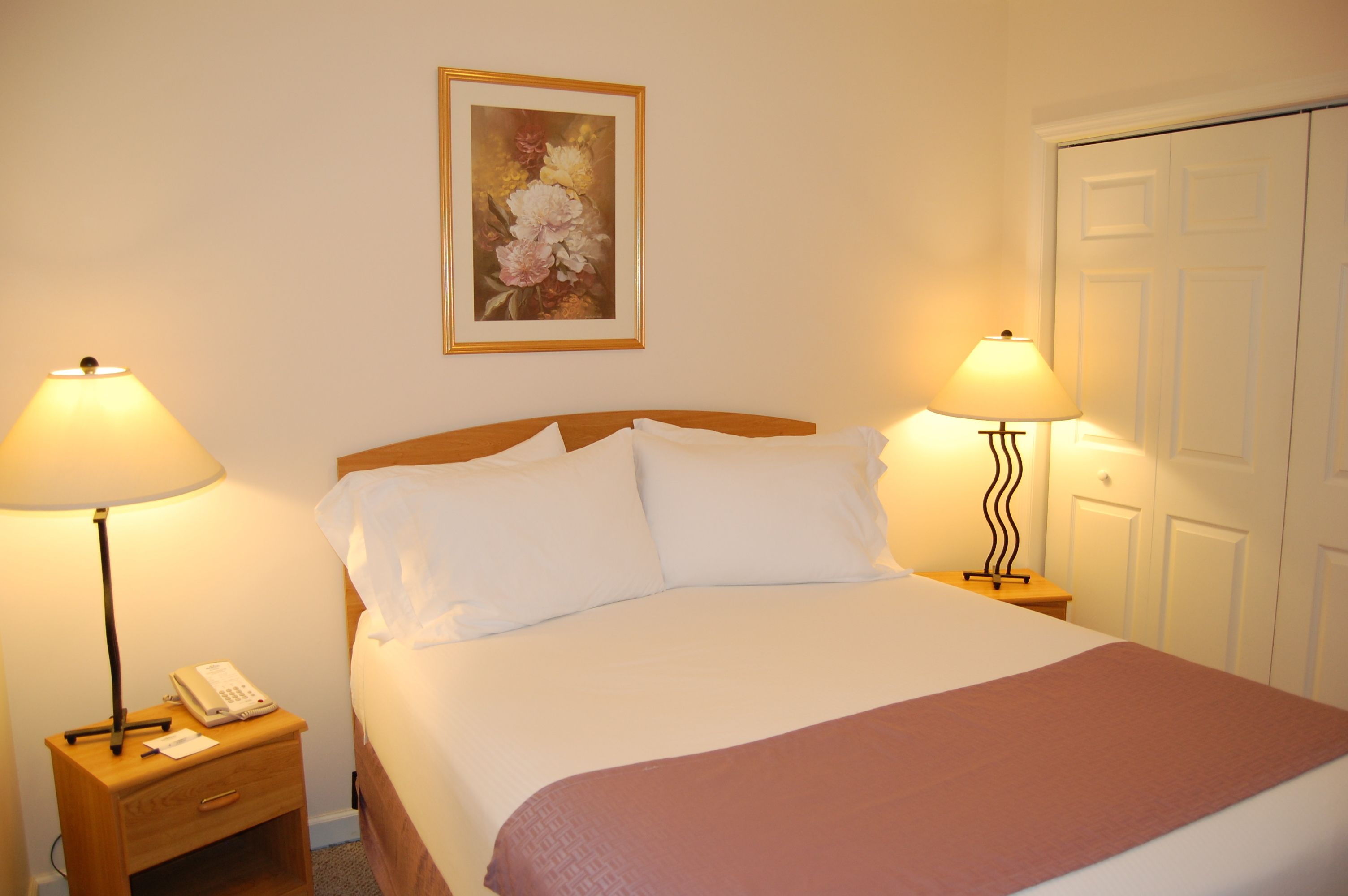 A neatly made bed with white linens and a pink throw, flanked by two bedside tables with lamps and a phone.