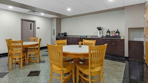 Una sala de conferencias con una mesa redonda y sillas de madera, una kitchenette con electrodomésticos y una puerta.