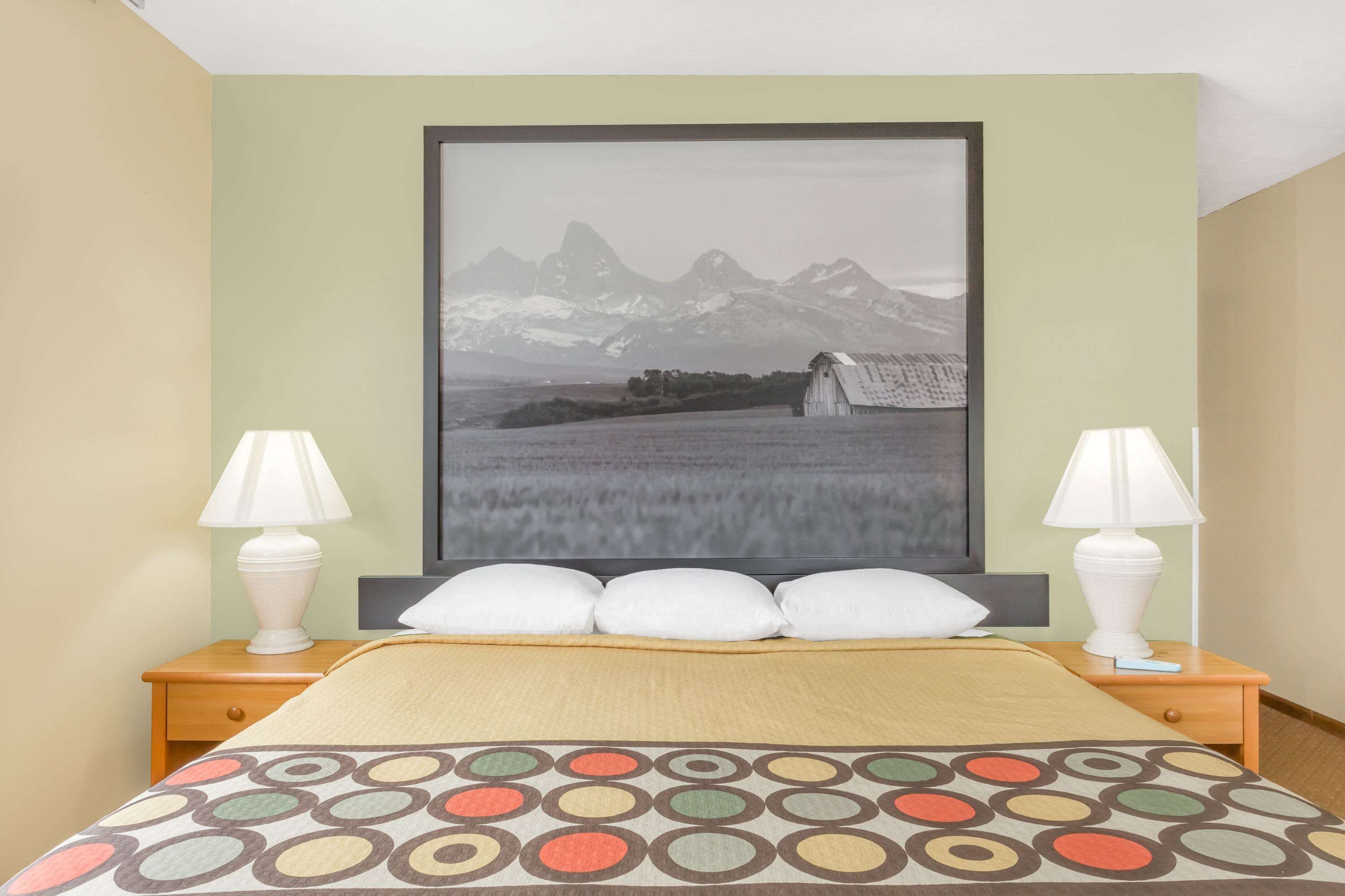A neatly made bed with a patterned bedspread, two bedside lamps, and a framed picture of a mountain landscape on the wall.