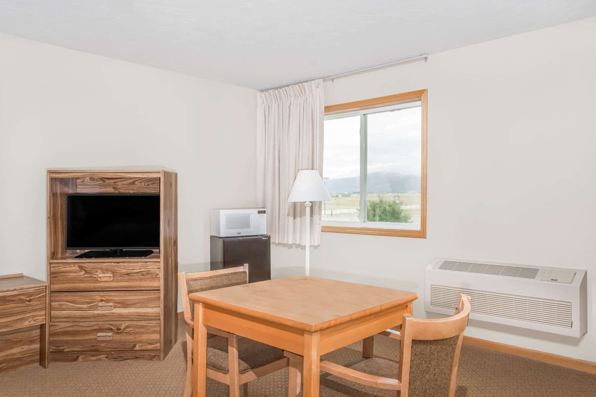 Een kamer met een houten tv-meubel, een kleine tafel met stoelen, een lamp, een magnetron en een raam met uitzicht op de omgeving.