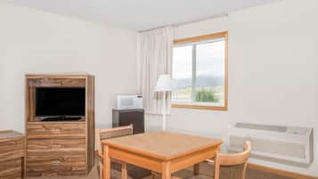 A room with a wooden TV stand, a small table with chairs, a lamp, a microwave, and a window with a view of the outdoors.