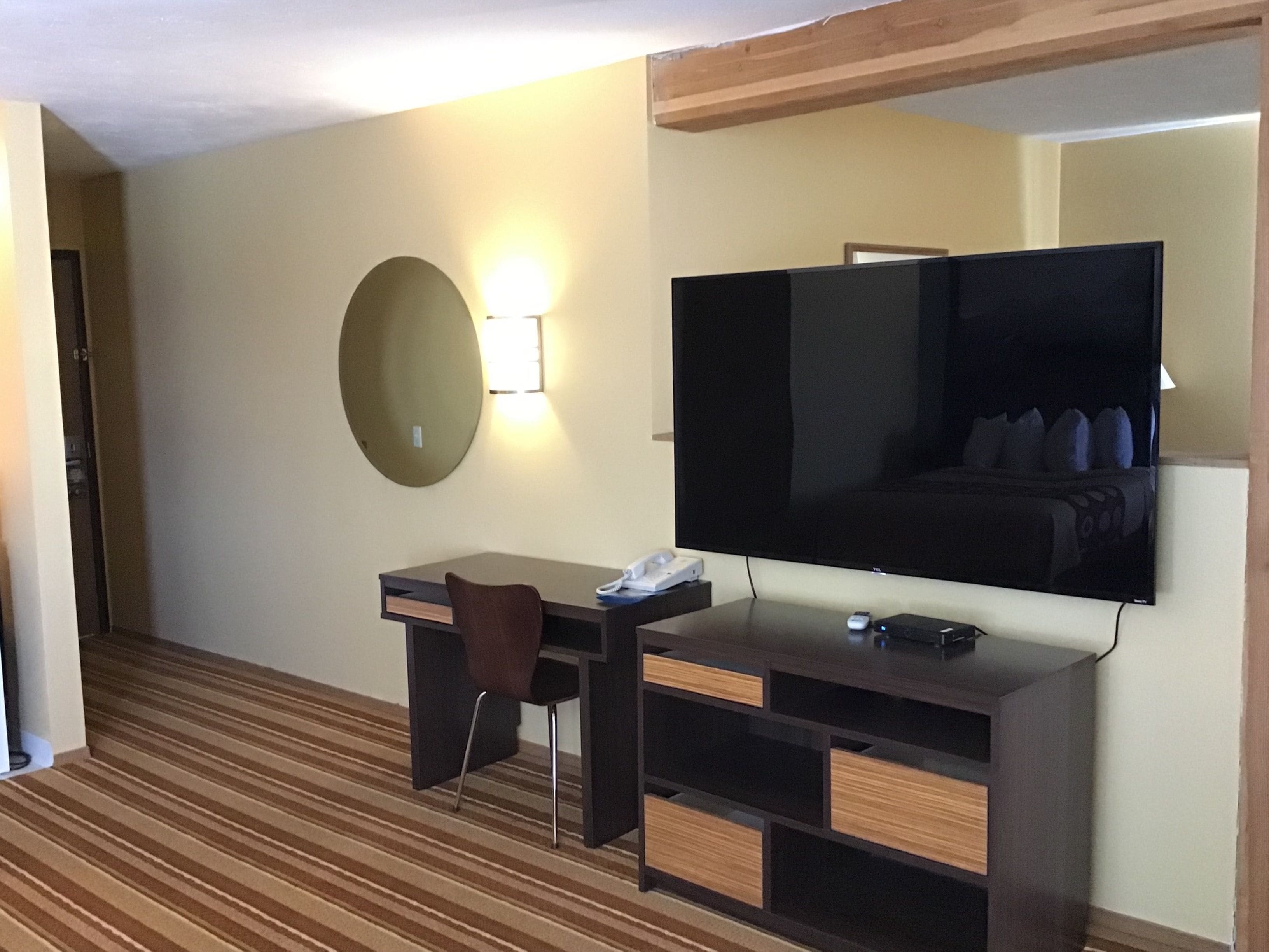 A hotel room with a large flat-screen TV mounted on the wall, a desk with a chair, a small bedside table, and a striped carpet.