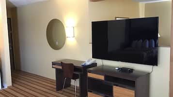 Habitación de hotel con televisor de pantalla plana grande montado en la pared, un escritorio con silla, una mesita de noche pequeña y alfombra a rayas.