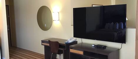 A hotel room with a large flat-screen TV mounted on the wall, a desk with a chair, a small bedside table, and a striped carpet.