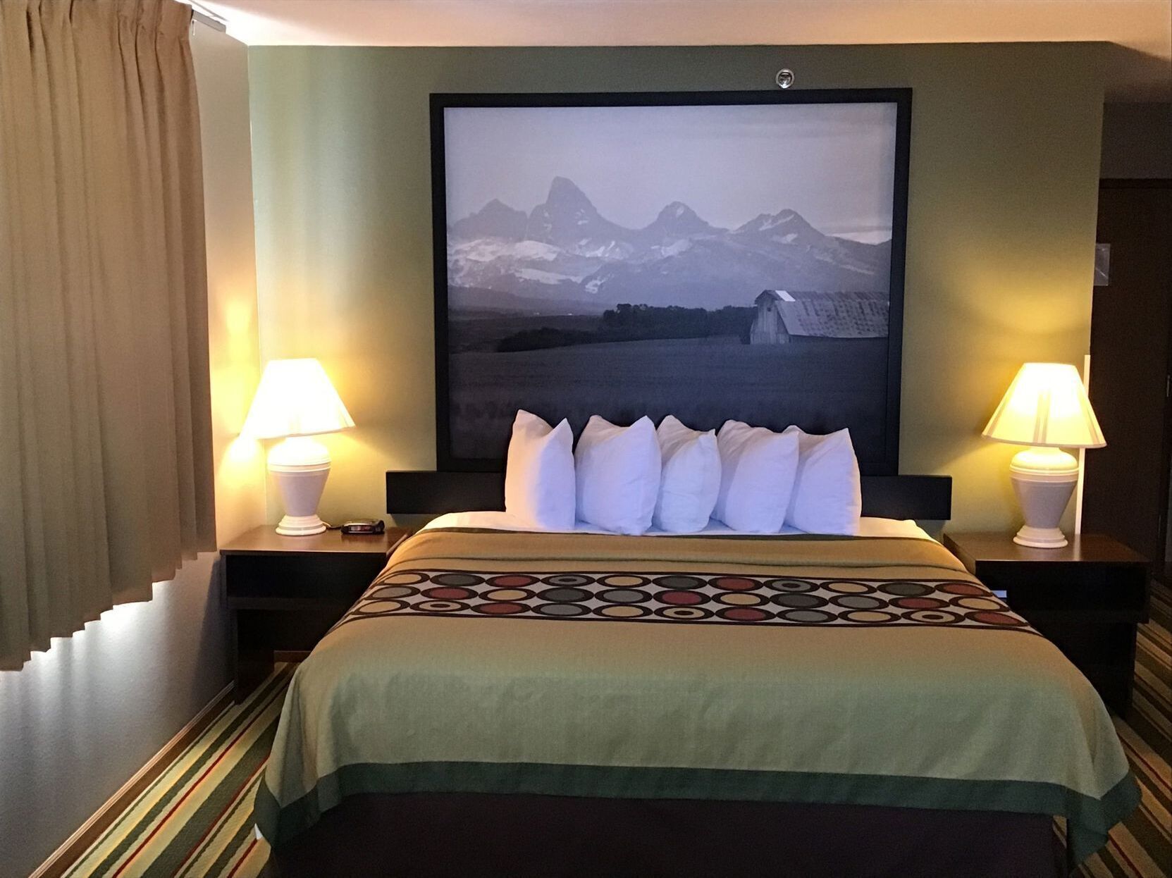 Een hotelkamer met een bed, twee bedlampen, een groot gepresenteerd schilderij aan de muur, en een gestreept tapijt.