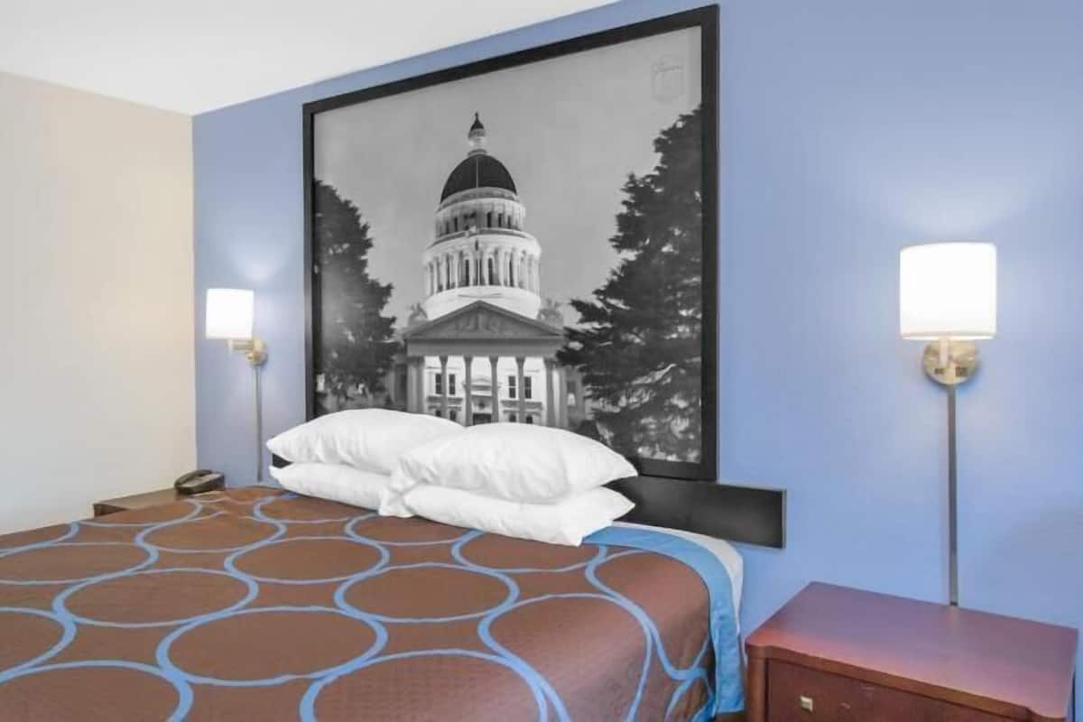 Een hotelkamer met een bed, nachtkastje, lamp en een aan de muur hangend schilderij van een gebouw.
