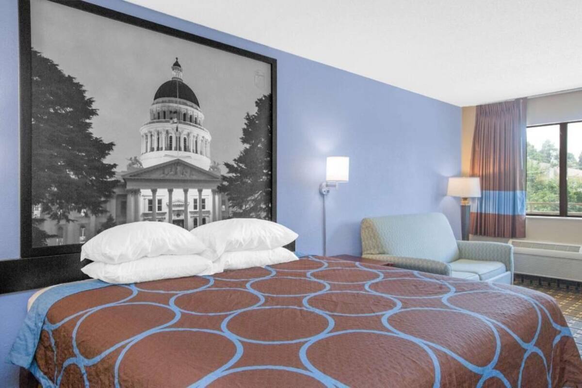 Een hotelkamer met een bed, een stoel, een lamp en een lijst met een afbeelding van een gebouw.
