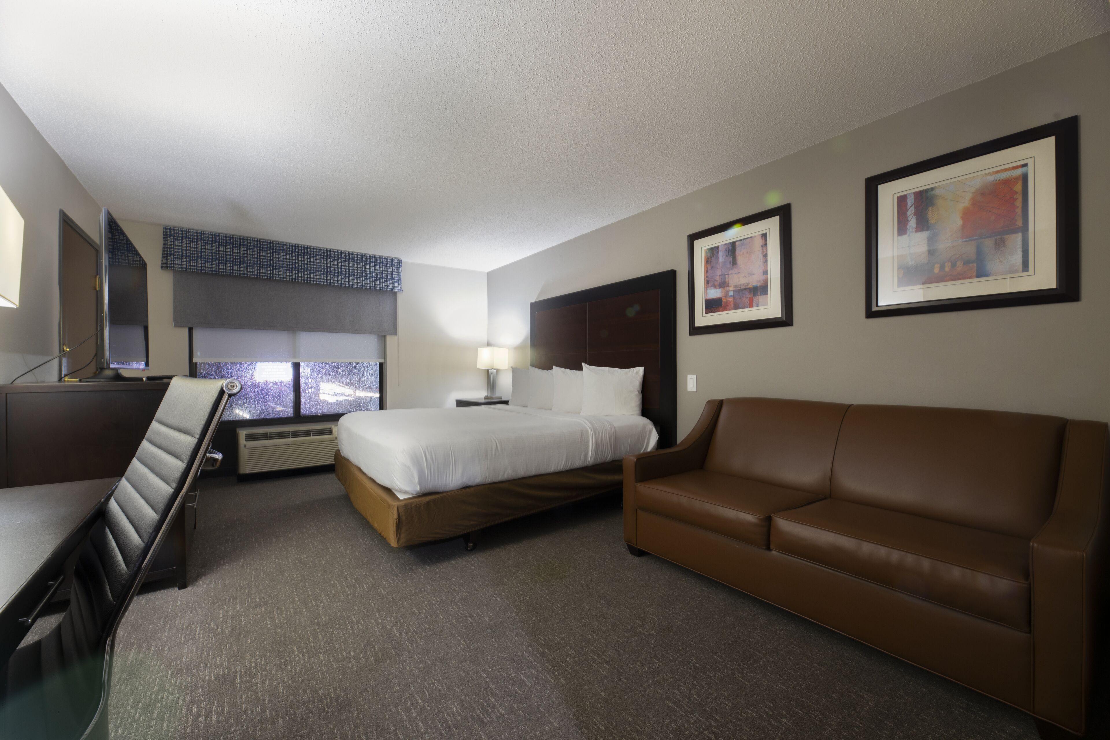 A hotel room with a bed, a brown leather sofa, a desk, and a window with blinds.