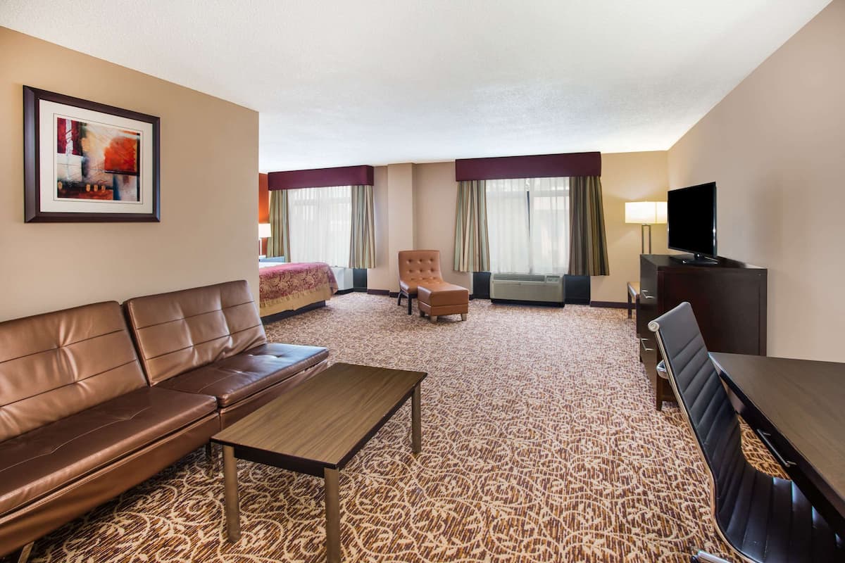 Une chambre d’hôtel avec un canapé en cuir marron, une table basse en bois, un petit fauteuil, un téléviseur posé sur un meuble et un tapis à motifs.