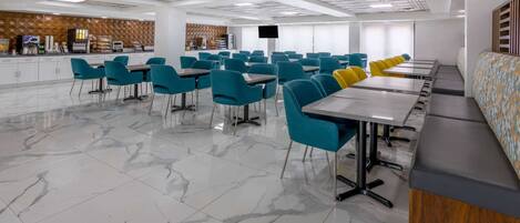 Une cafétéria spacieuse avec un sol en marbre, des chaises turquoise et jaunes, et un long comptoir doté d’un espace dédié au café.