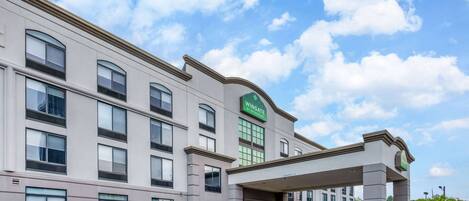 A multi-story hotel with a modern facade, a prominent green sign, and a covered entrance.