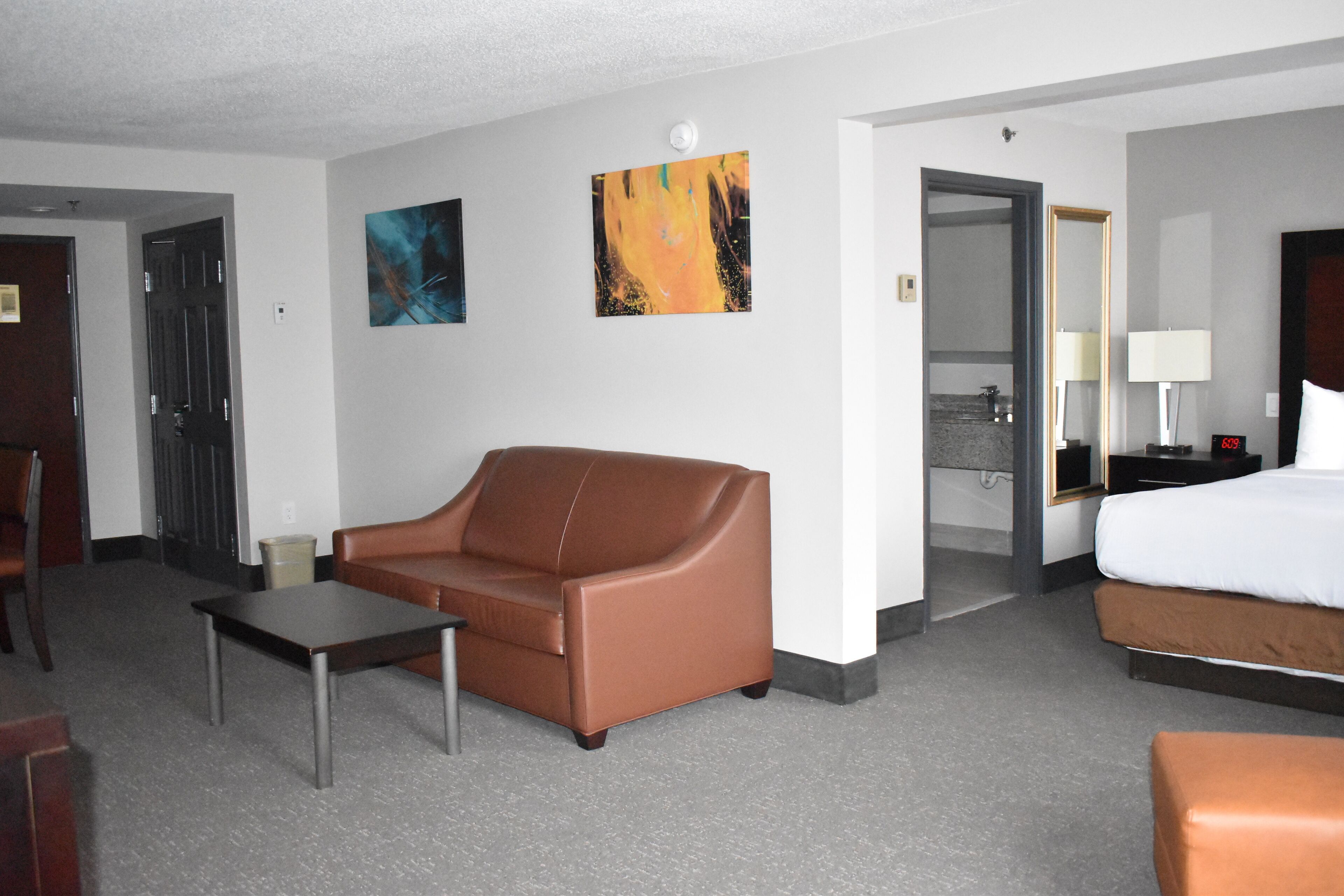 Habitación de hotel con un sofá de cuero marrón, una mesita, una cama con ropa de cama blanca, una mesita de noche con lámpara y dos cuadros en la pared.
