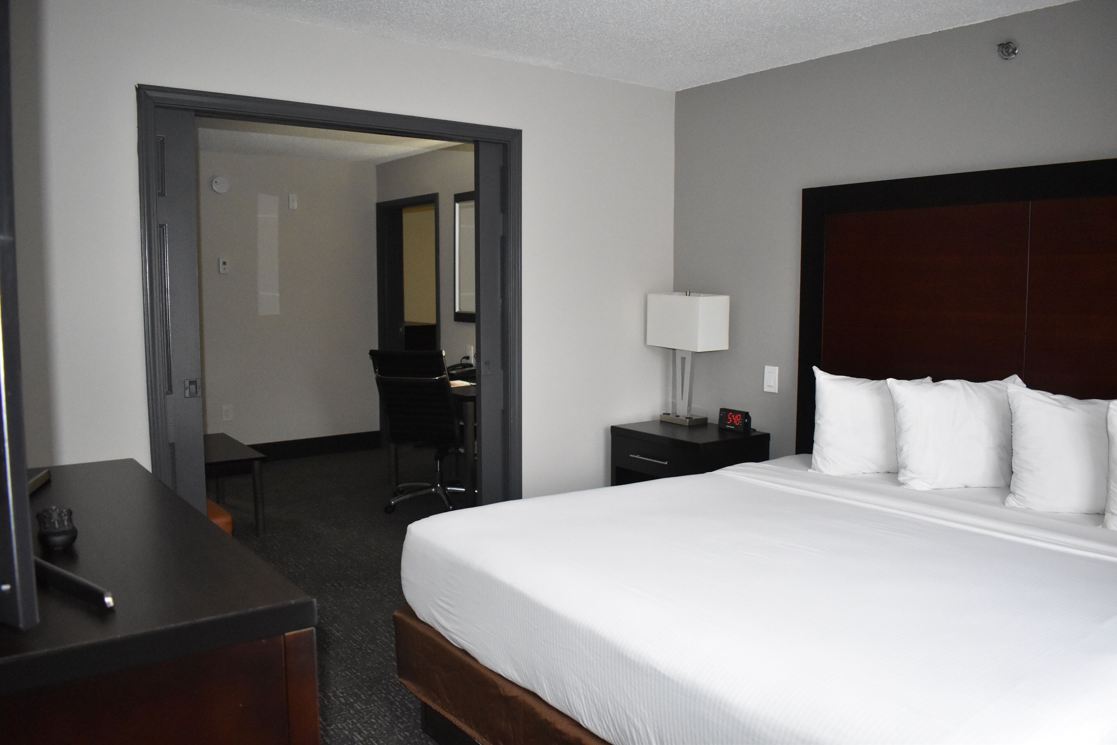 A hotel room with a large bed, a desk, a chair, and a nightstand with a lamp.