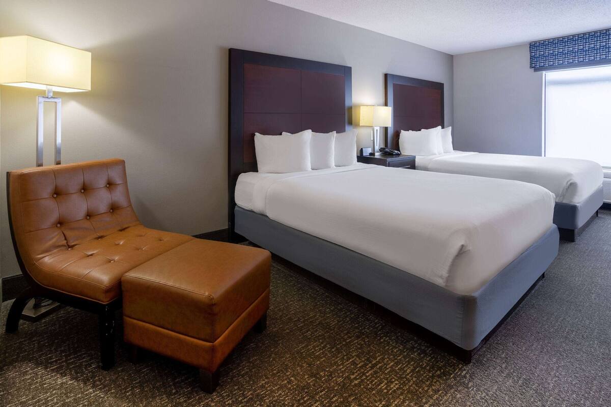 Une chambre d’hôtel avec un grand lit, un lit plus petit, un fauteuil en cuir marron et une table de chevet avec une lampe.