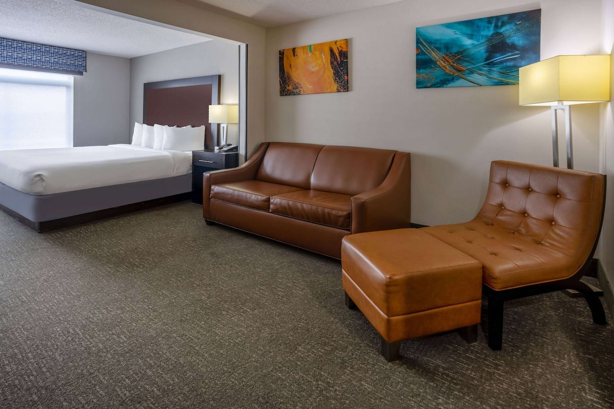 Une chambre d’hôtel avec un lit, un canapé en cuir marron, un pouf assorti et un fauteuil. Il y a deux tableaux au mur et une lampe sur une table d’appoint.
