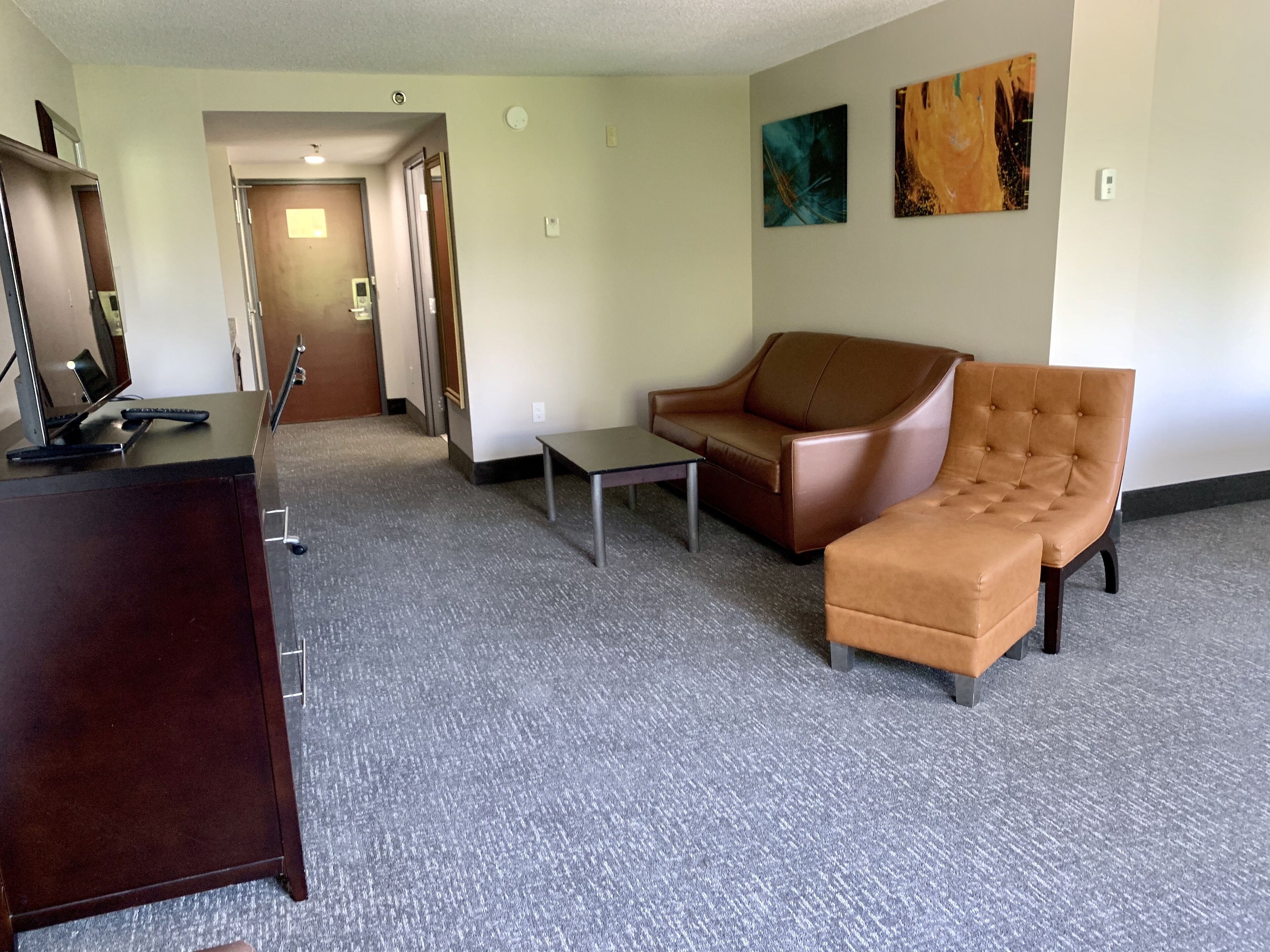 A hotel room with a desk, a brown leather sofa, a small table, and two paintings on the wall.