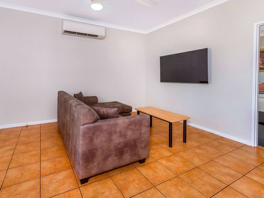 A living room with a brown sofa, a small wooden table, a flat-screen TV mounted on the wall, and an air conditioning unit.