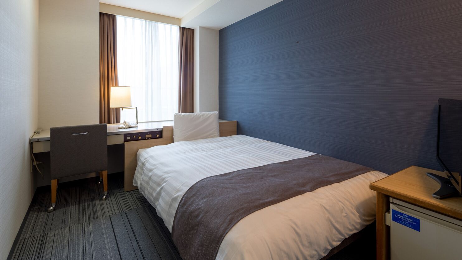 Quarto de hotel com cama, escrivaninha, cadeira, abajur e televisão.