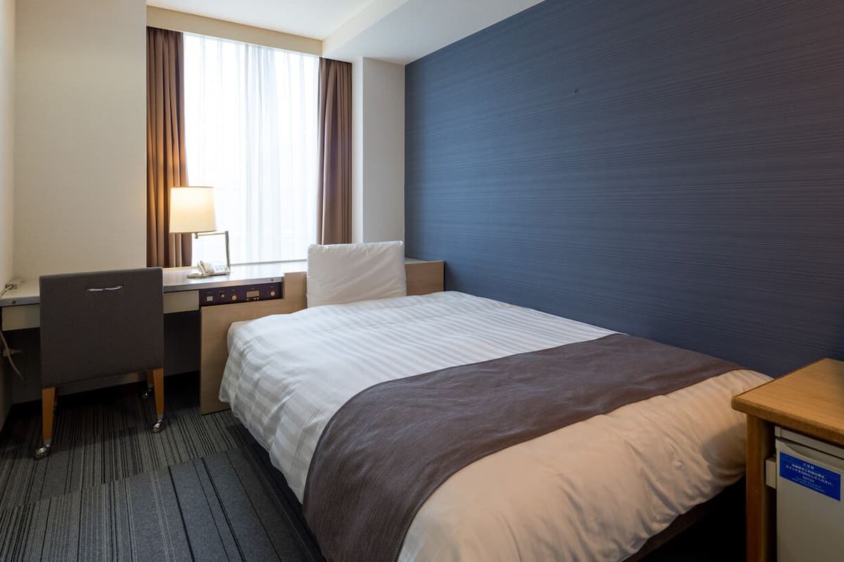 A hotel room with a bed, desk, chair, lamp, and TV.