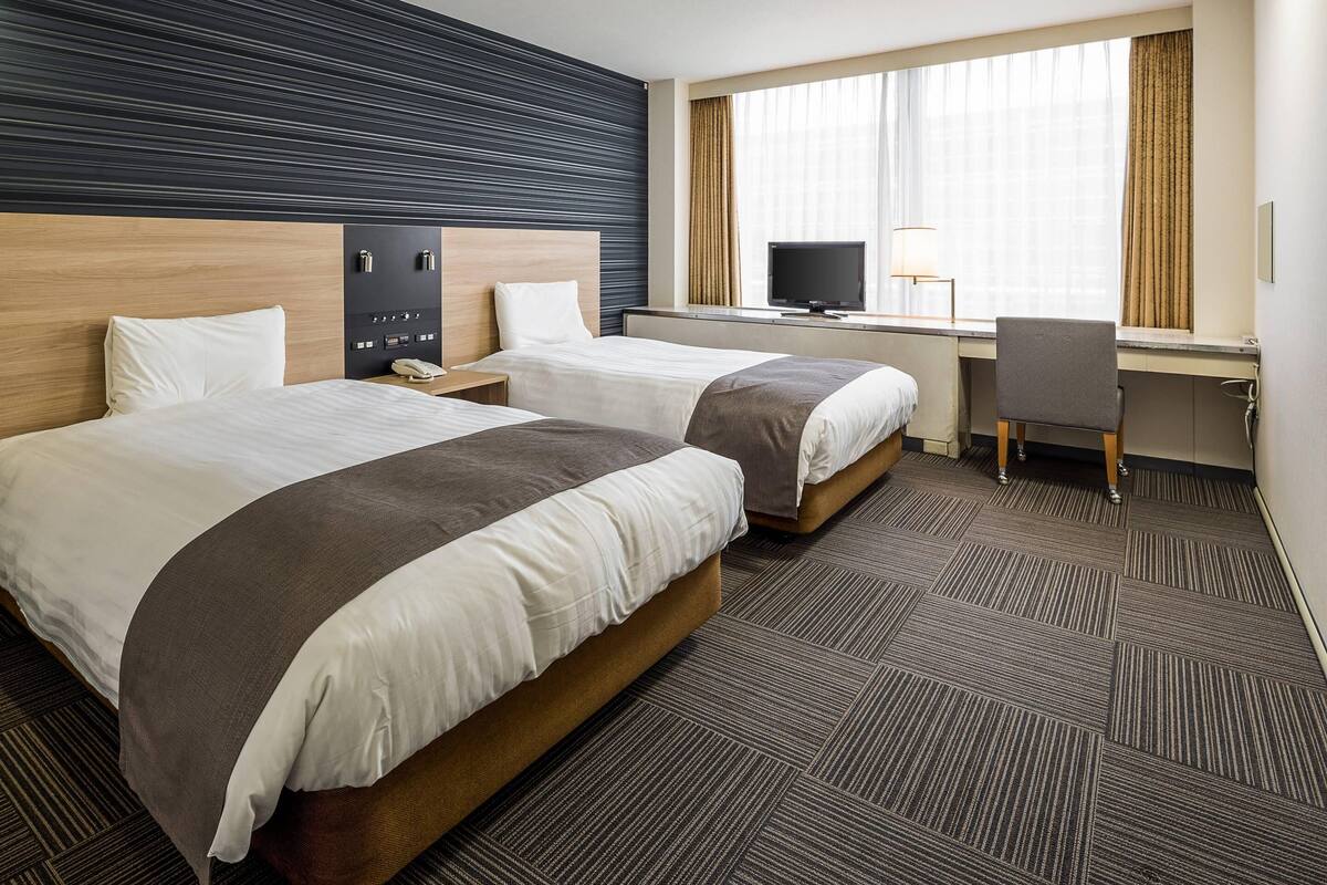A hotel room with two beds, a desk, a chair, a TV, and a window with curtains.