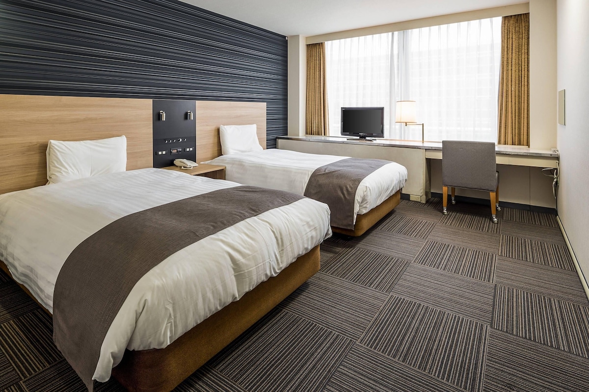 A hotel room with two beds, a desk, a chair, a TV, and a window with curtains.