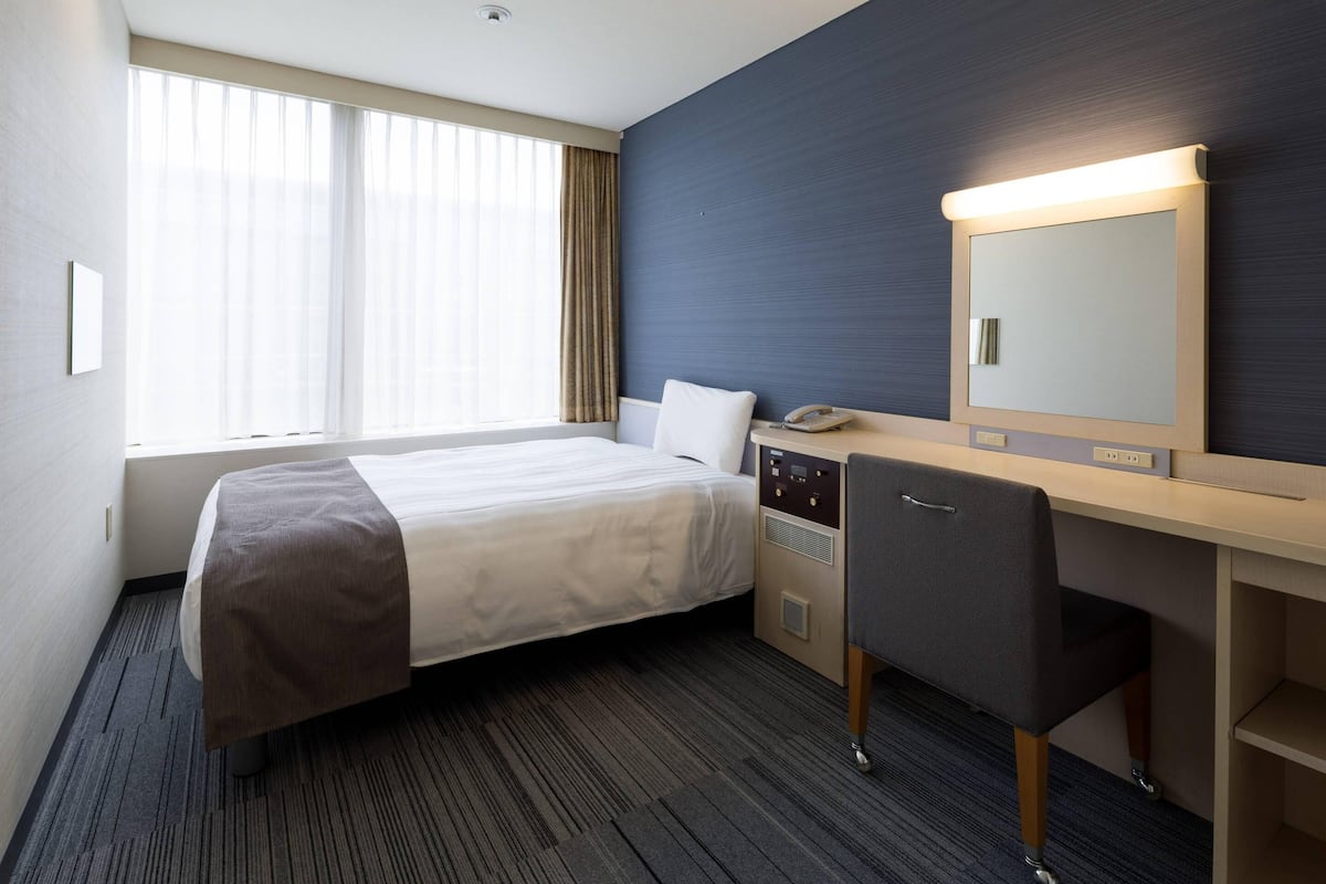 A hotel room with a bed, a desk with a chair, a mirror, and a window with curtains.