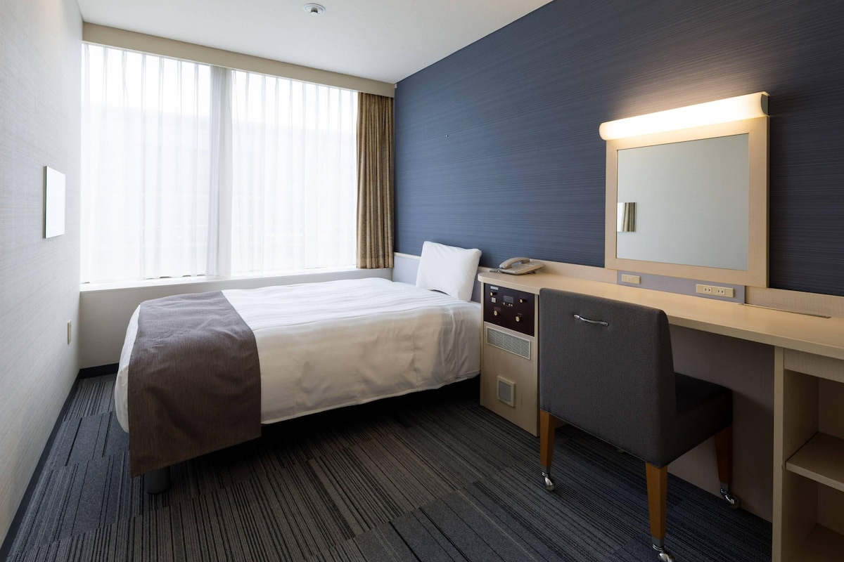 A hotel room with a bed, a desk with a chair, a mirror, and a window with curtains.