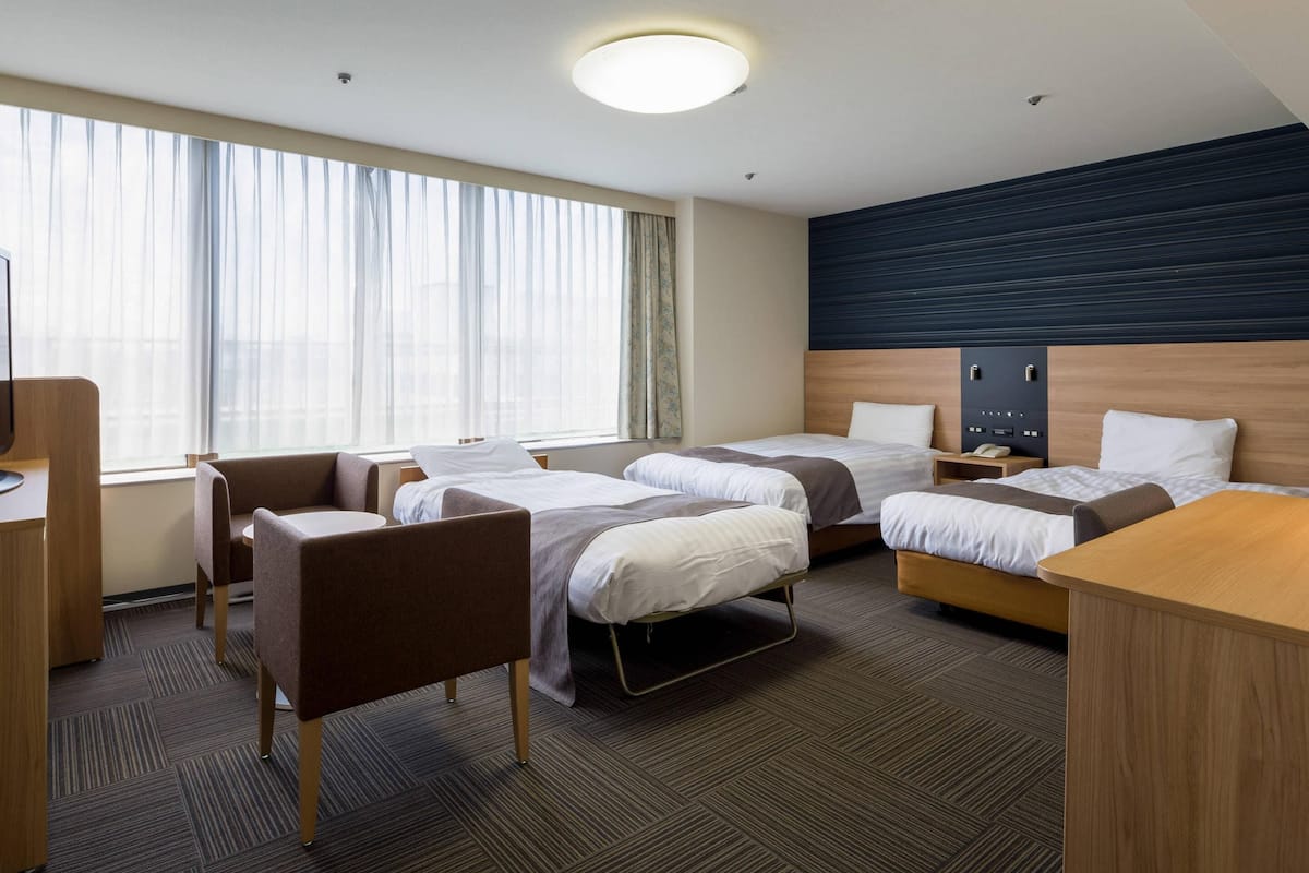 A hotel room with two beds, a desk, a chair, a window with curtains, and a headboard with a control panel.