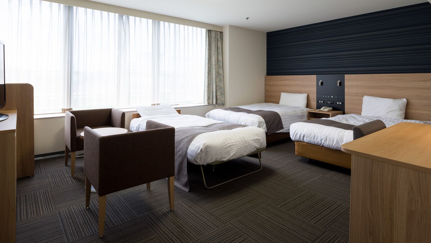 Quarto de hotel com duas camas, uma escrivaninha, uma cadeira e uma janela grande.