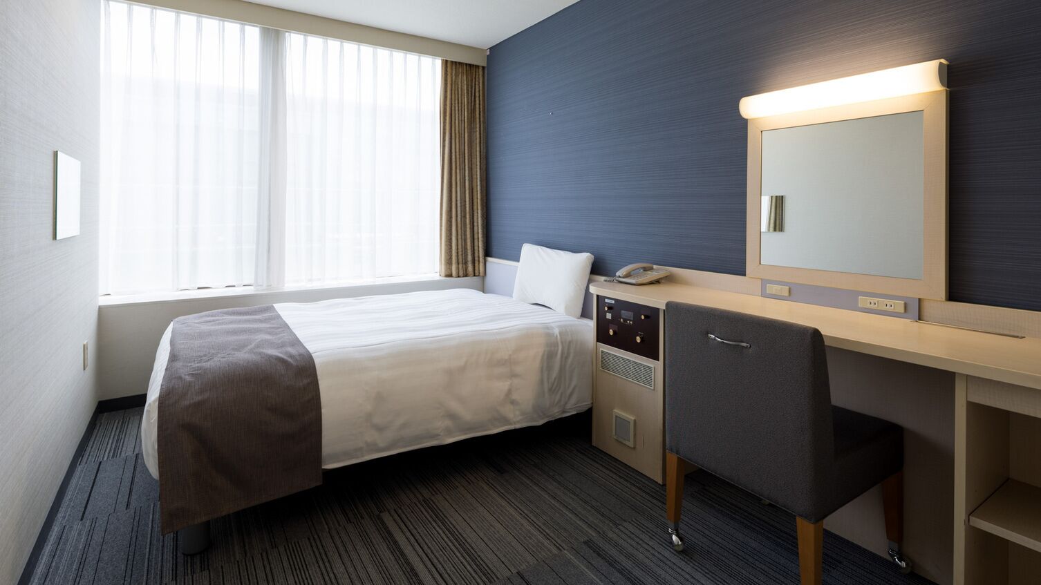 Quarto de hotel com cama, escrivaninha, cadeira, telefone e um espelho grande.