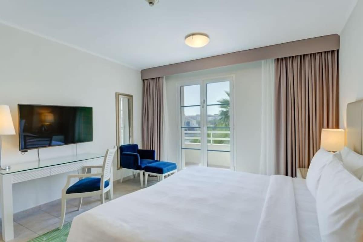 A hotel room with a bed, a desk with a chair, a television, and a window with curtains.
